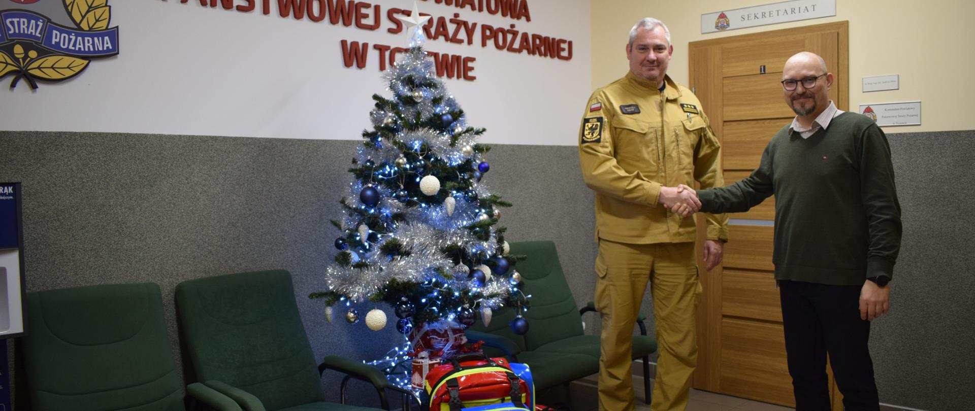 Komendant uściskuje rękę przedstawiciela firmy Huber+Suhner. Obok fotele, choinka i pod choinką przekazany sprzęt ratownictwa medycznego, plecak i inne torby. W tle wejście do sekretariatu.