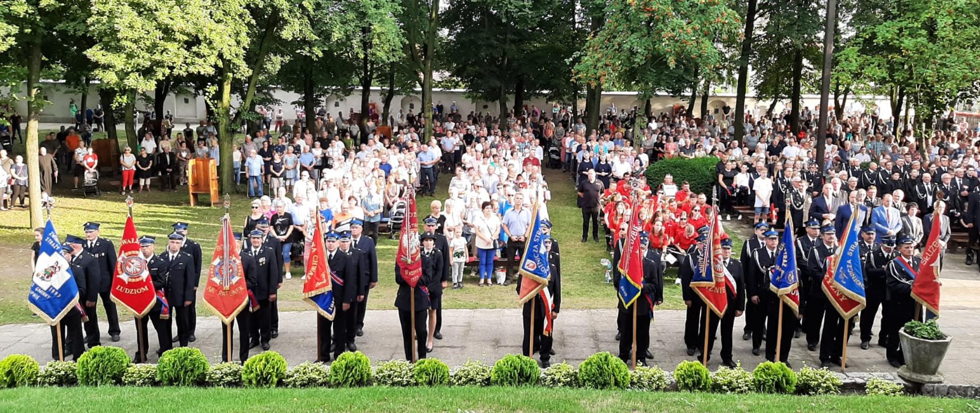 Strażacy ze sztandarami stoją w szeregu przed sanktuarium w Górce Duchownej. W tle widoczne stojące osoby cywilne.