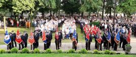 Strażacy ze sztandarami stoją w szeregu przed sanktuarium w Górce Duchownej. W tle widoczne stojące osoby cywilne.