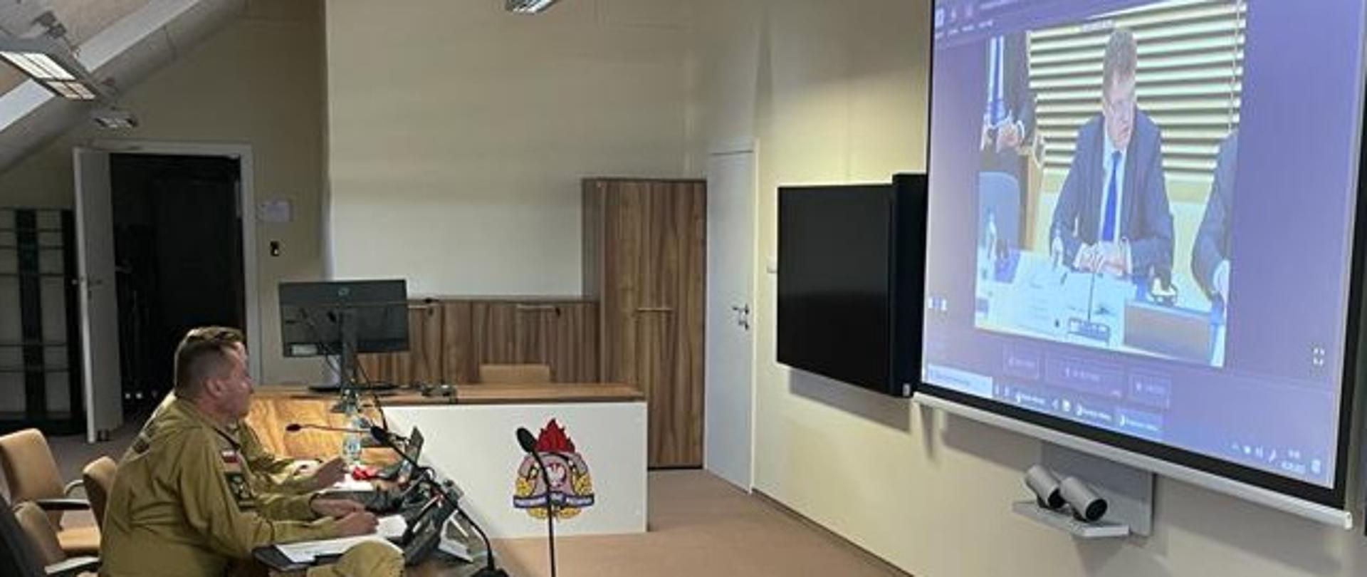 Zastępca komendanta głównego PSP wraz z zastępcą dyrektora biura planowania operacyjnego KG PSP siedzą przy biurku podczas uczestnictwa w formie wideokonferencji w szczycie zorganizowanym w trybie hybrydowym w Brukseli. Przed nimi na ścianie monitor ekranowy z uczestnikami wideospotkania