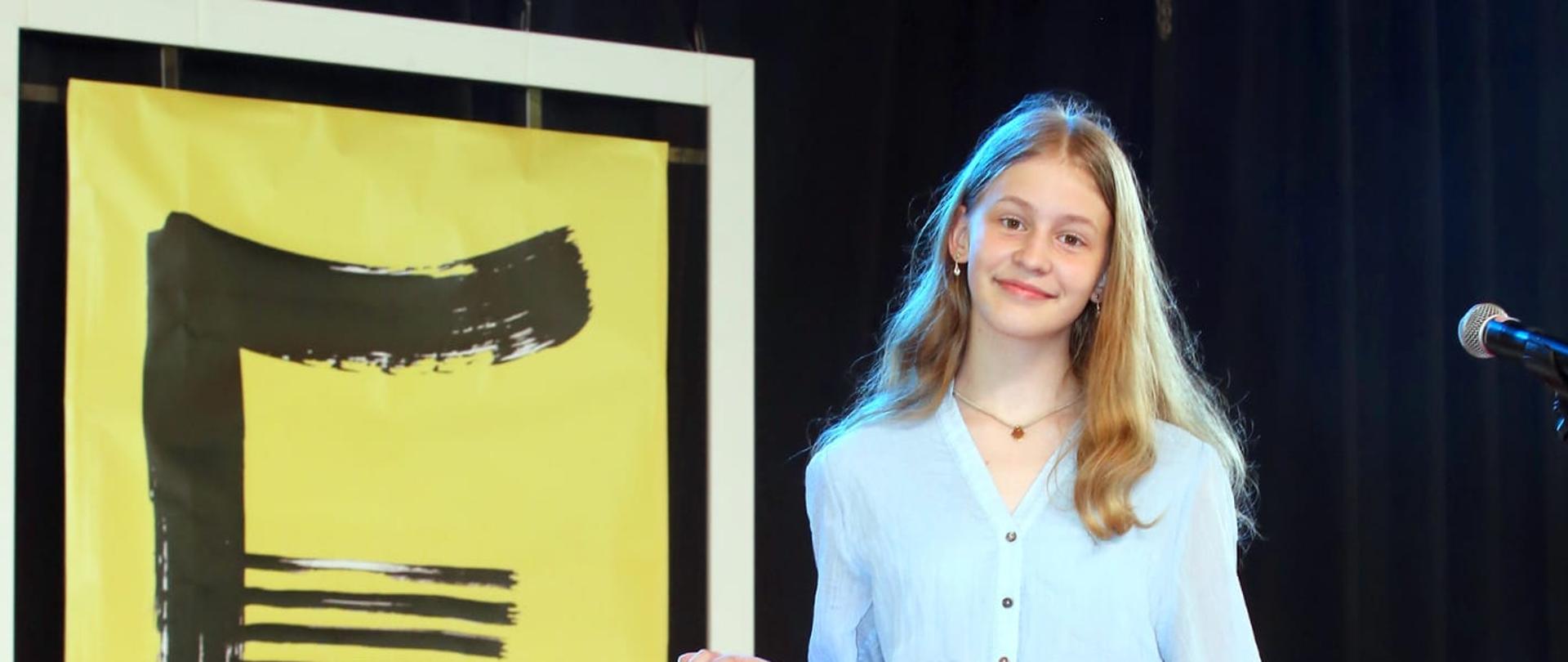 Dziewczyna ubranw niebiską sukienkę stoj.ąca na scenie. W tle żółto-czarny plakat konkursu.