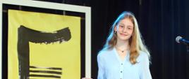Dziewczyna ubranw niebiską sukienkę stoj.ąca na scenie. W tle żółto-czarny plakat konkursu.