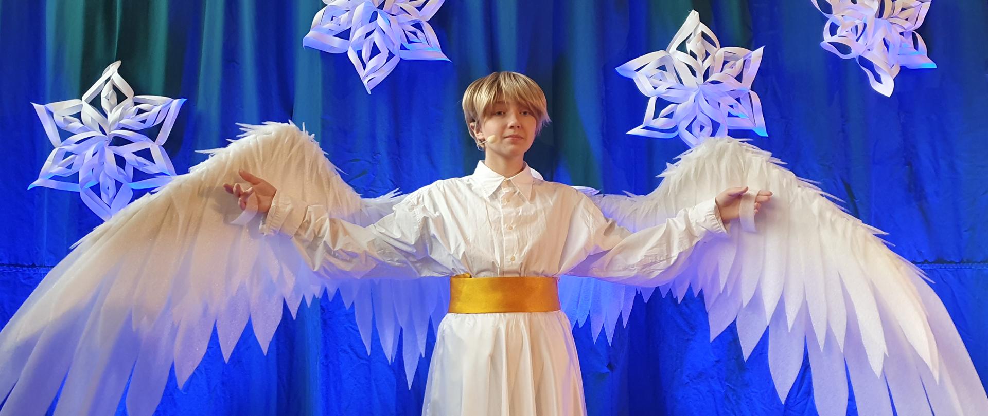 Na zdjęciu w części centralnej znajduje się uczennica w stroju anioła. Postać ubrana jest w białą suknię i jest przepasana złotym pasem. Postać ma lekko uniesione ręce do góry. Do rąk anioła przyczepione są białe rozpostarte skrzydła. Postać anioła biorącego udział w Jasełkach przedstawiona jest na błękitnym tle. Nad postacią zawieszone są cztery gwiazdy, wykonane z białego kartonu.