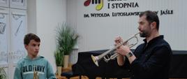 Mężczyzna w czarnej koszuli gra na trąbce. Z lewej strony stoi chłopiec trzymający w ręku trąbkę i patrzy na grającego mężczyznę. W tle stoi czarny fortepian, a za nim zielone kwiaty. W tle ściana w jasnym kolorze z napisem państwowa szkoła muzyczna pierwszego stopnia im Witolda Lutosławskiego w Jaśle obok napisu logo szkoły przedstawiające dwie kolorowe nutki, między którymi widać trzy klawisze fortepianu.