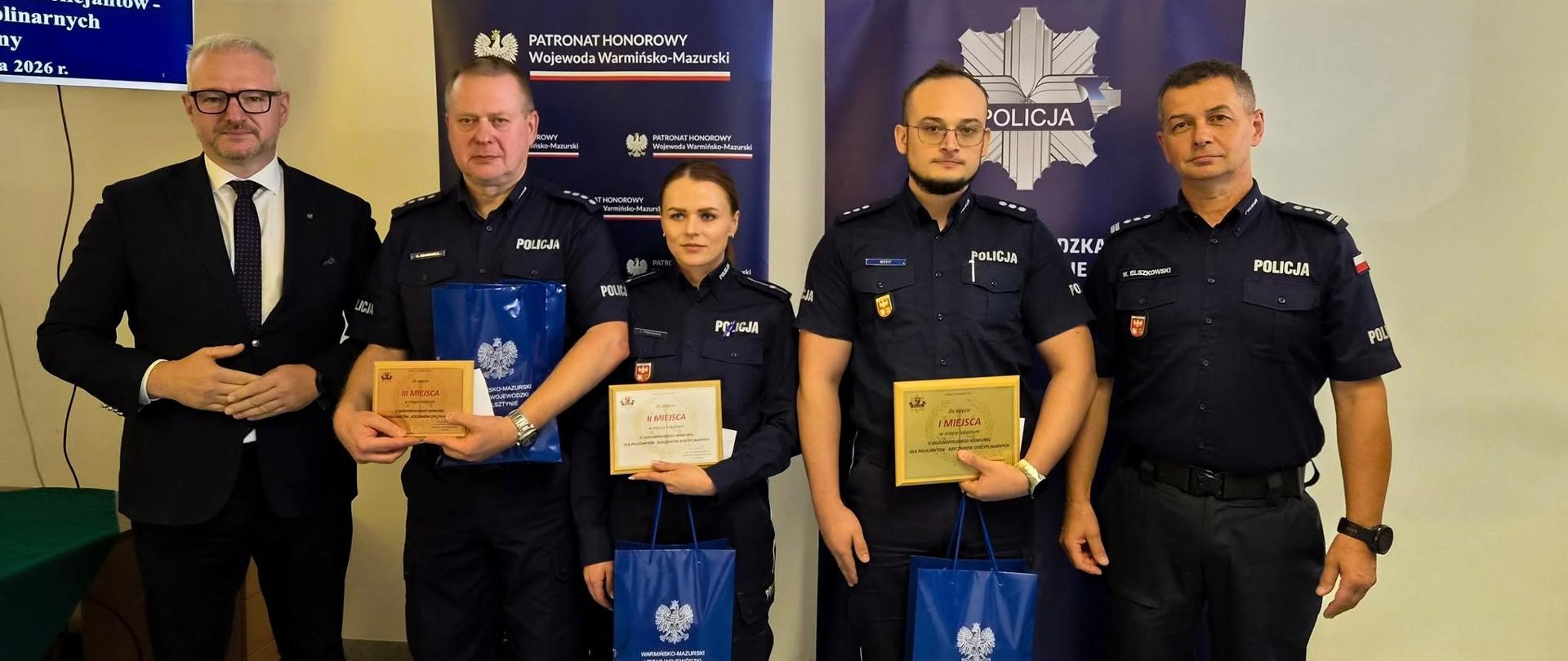 Grupa osób, w tym policjanci w mundurach oraz wojewoda warmińsko-mazurski w garniturze, pozuje do zdjęcia. Trzech policjantów trzyma wyróżnienia, tabliczki oraz torby z upominkami z logo Wojewody.