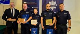 Grupa osób, w tym policjanci w mundurach oraz wojewoda warmińsko-mazurski w garniturze, pozuje do zdjęcia. Trzech policjantów trzyma wyróżnienia, tabliczki oraz torby z upominkami z logo Wojewody.