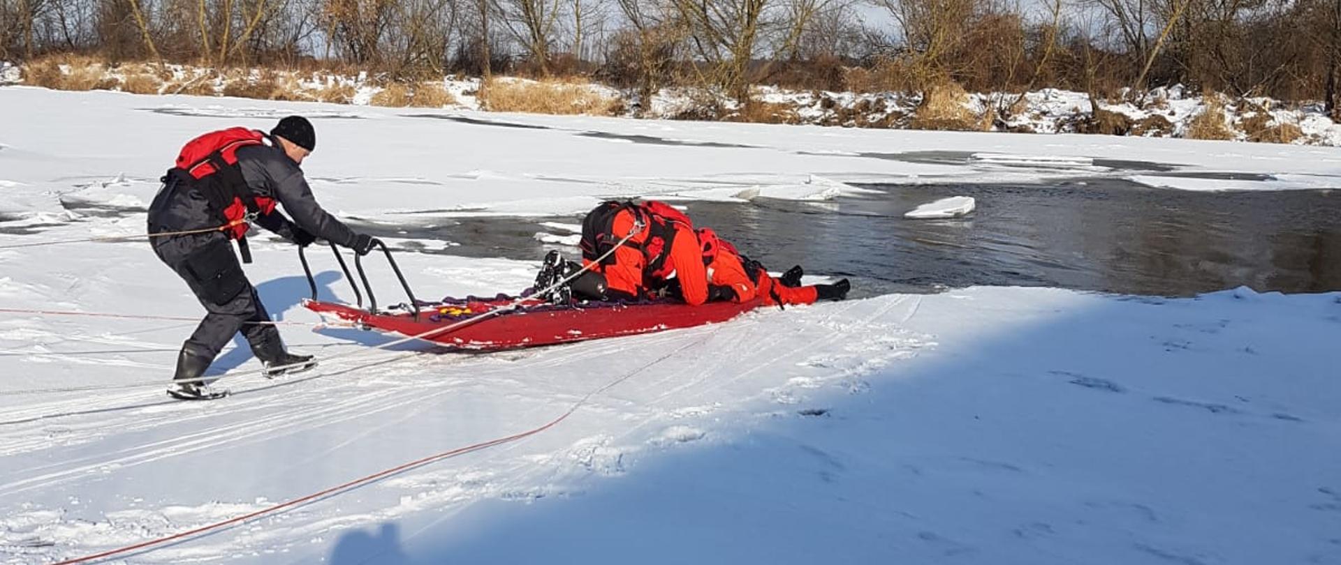 Dwóch ratowników na zamarzniętym akwenie przy pomocy sań lodowych podejmuje próbę wydobycia poszkodowanego na brzeg.