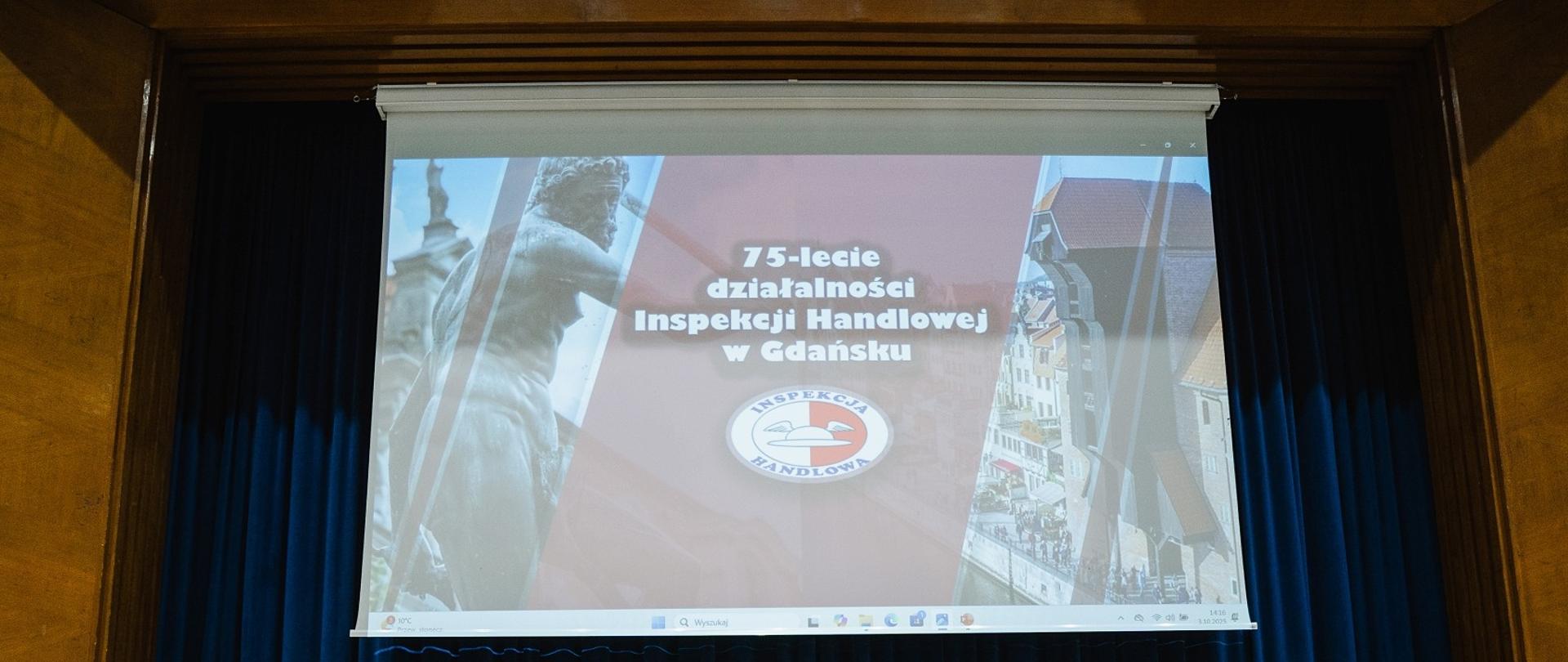 Telebim z wyświetlonym napisem 75-lecie działalności Inspekcji Handlowej w Gdańsku w tle granatowa kurtyna.