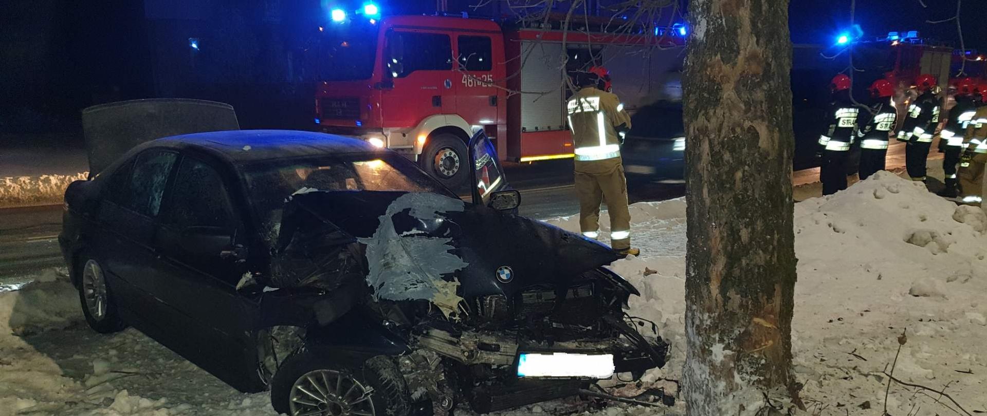Na zdjęciu widać samochód osobowy BMW oraz drzewo w które uderzył. Samochód posiada uszkodzony przód. W tle widać samochody straży pożarnej z włączona sygnalizacja świetlną, oraz ratowników którzy usuwają skutki zdarzenia. 