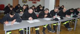 Egzamin końcowy. Świetlica Komendy Powiatowej PSP w Rawiczu. Na tle ściany z nazwą komendy i logiem PSP przy stołach ustawionych w 3 rzędach siedzą kursanci, którzy rozwiązują test pisemny. Strażacy są ubrani w "koszarówki". 