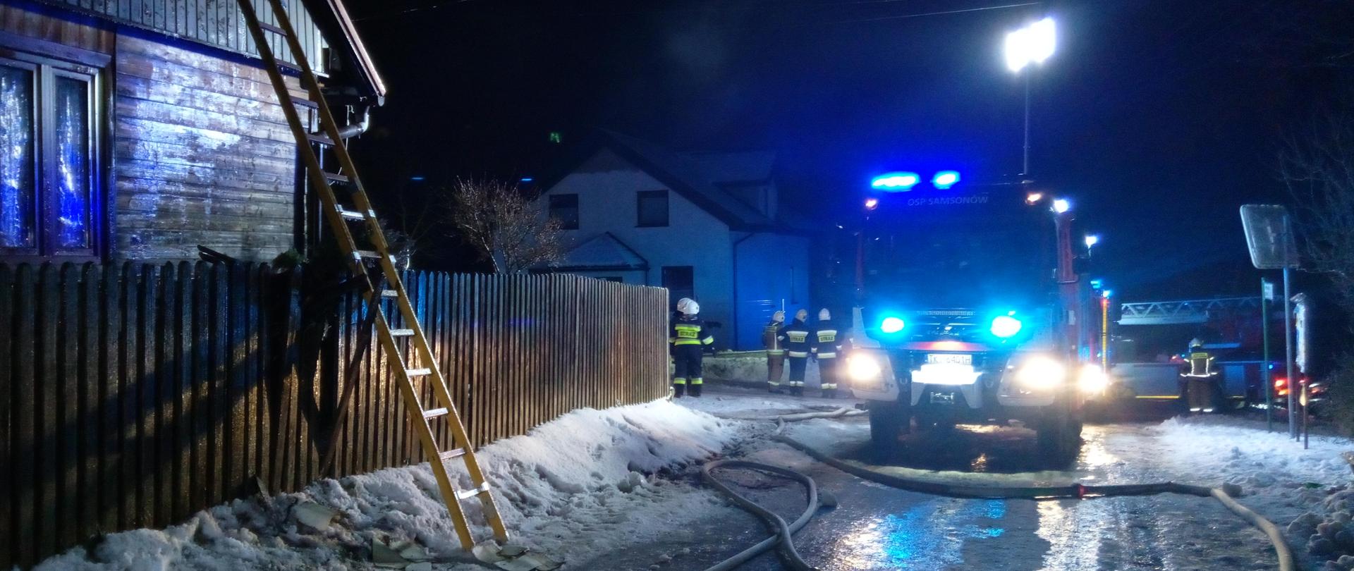 Zdjęcie przedstawia teren akcji. Po lewej dom do którego dostawiona jest drabina. Po prawej stoi samochód straży pożarnej. W oddali widać strażaków, przed samochodem rozwinięte linie gaśnicze. Nie widać dymu i ognia. Jest ciemno.