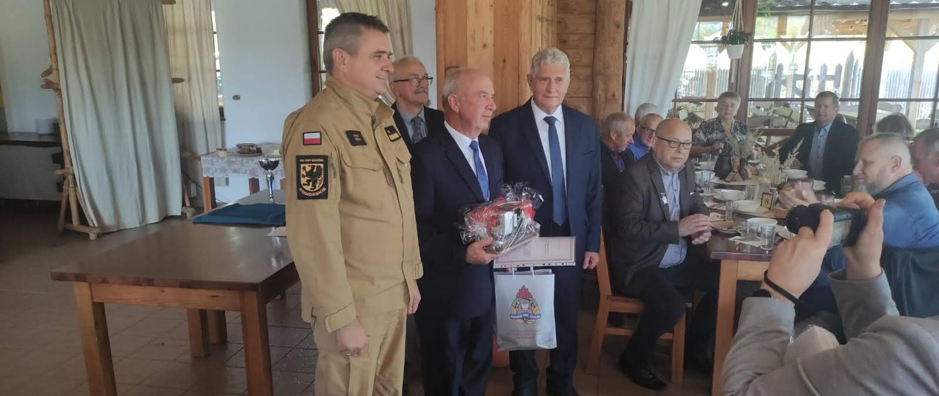 Pomorski komendant wojewódzki Państwowej Straży Pożarnej stoi z wyróżnionym mężczyzną obok stoją inni mężczyźni pozostali goście siedzą za stołem.