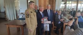 Pomorski komendant wojewódzki Państwowej Straży Pożarnej stoi z wyróżnionym mężczyzną obok stoją inni mężczyźni pozostali goście siedzą za stołem.
