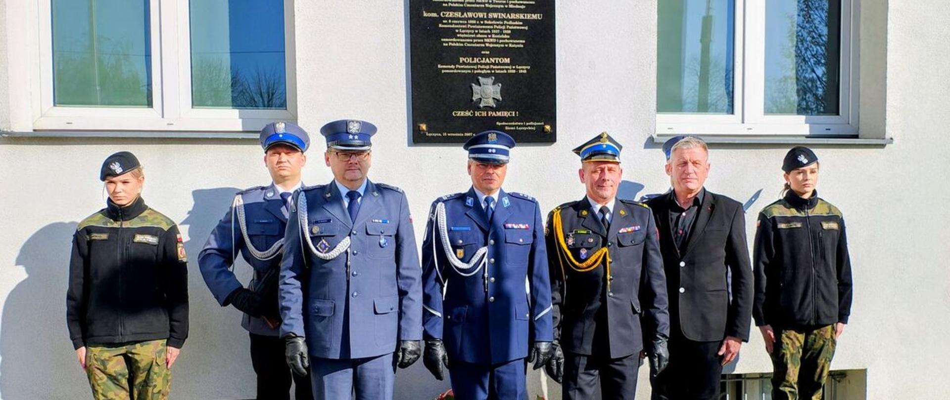 Na zdjęciu widoczne są obchody Dnia Pamięci Ofiar Zbrodni Katyńskiej. Przed tablicą pamiątkową stoją w szeregu funkcjonariusze w galowych mundurach, żołnierze oraz mężczyzna w stroju cywilnym. Uroczystość ma charakter oficjalny i podniosły — uczestnicy stoją na baczność, oddając hołd ofiarom. Pod tablicą złożono kwiaty.