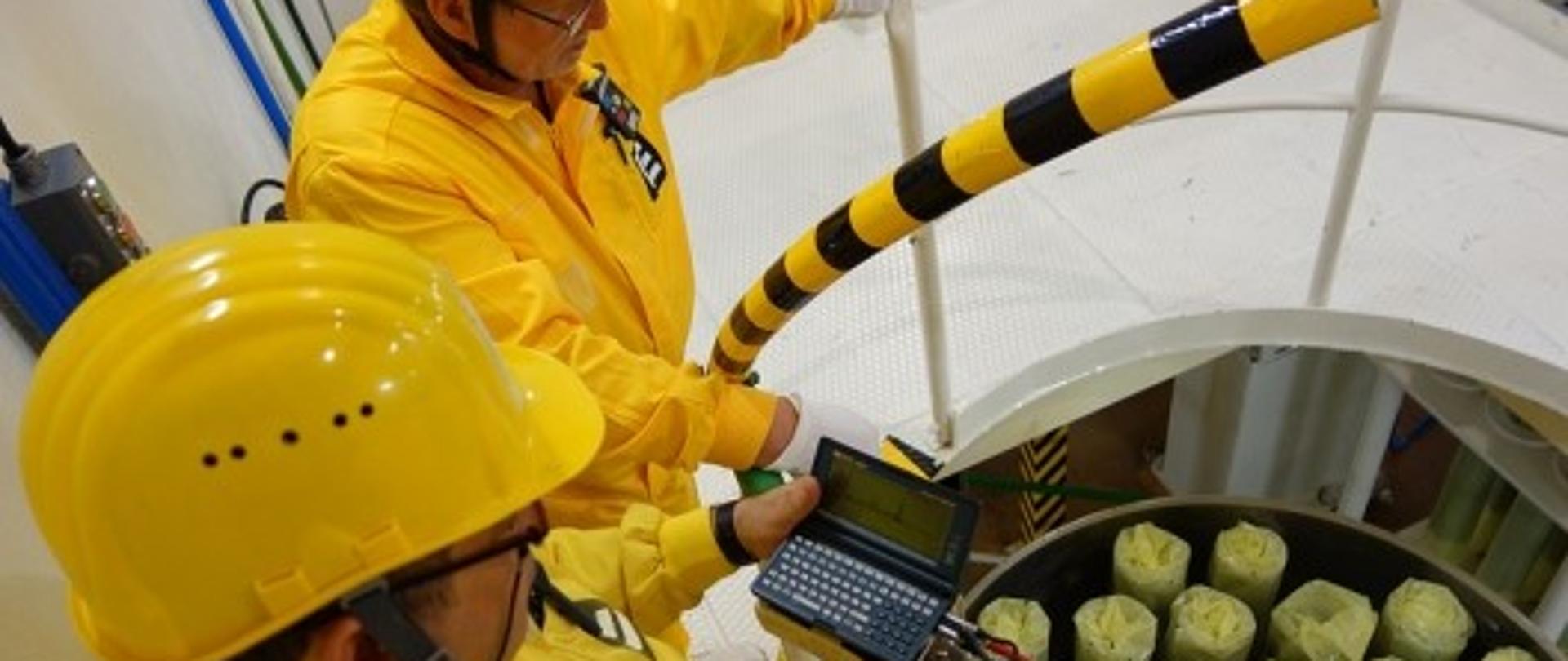 Zdjęcie przedstawia 2 mężczyzn w żółtych kombinezonach ochronnych w żółtych kaskach z przyrządem mierniczym kontrolujących świeże paliwo jądrowe 