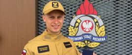 Zdjęcie przedstawia uśmiechniętego strażaka w piaskowym mundurze, który stoi przed budynkiem Komendy Powiatowej Państwowej Straży Pożarnej w Garwolinie. Mężczyzna prezentuje trzymaną w dłoni małą, niebieską legitymację z wyraźnym napisem „Dawca przeszczepu”. Na jego umundurowaniu widoczny jest imiennik „Kędziorek Mateusz”, a w tle umieszczono duże godło oraz czerwoną tablicę informacyjną formacji.
