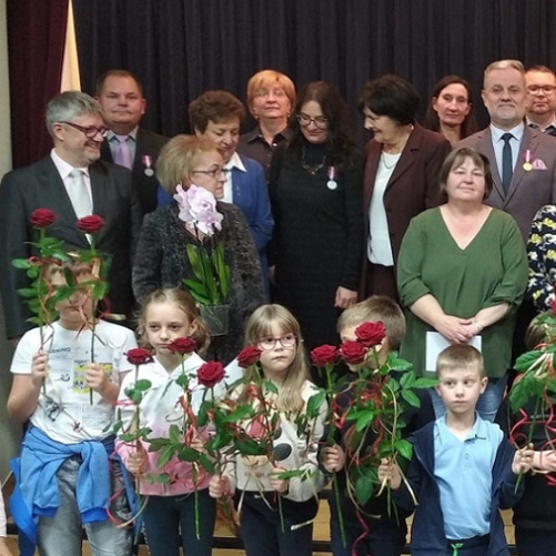 Zdjęcie przedstawia osoby ustawione w trzech rzędach na scenie w sali koncertowej szkoły. W pierwszym rzędzie stoją małe dzieci z różami czerwonymi w rękach. W drugim i trzecim rzędzie, na schodach sceny, stoją nauczyciele i pracownicy szkoły odświętnie ubrani w radosnych nastrojach.