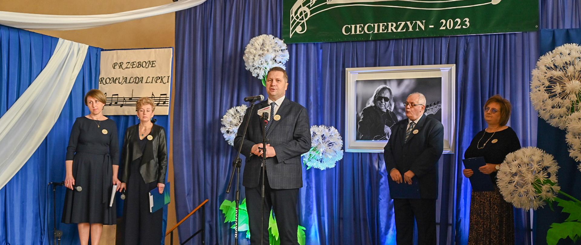 Minister Czarnek stoi i mówi do mikrofonu na stojaku, za nim żółta ściana na której wisi niebieska tkanina, a na niej czarno-białe zdjęcie kobiety z gitarą i zielona plansza z napisem XVI Powiatowy Festiwal Piosenki „Niezapomniane Melodie” Ciecierzyn 2023, obok stoi mężczyzna w garniturze i trzy kobiety.