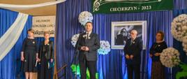 Minister Czarnek stoi i mówi do mikrofonu na stojaku, za nim żółta ściana na której wisi niebieska tkanina, a na niej czarno-białe zdjęcie kobiety z gitarą i zielona plansza z napisem XVI Powiatowy Festiwal Piosenki „Niezapomniane Melodie” Ciecierzyn 2023, obok stoi mężczyzna w garniturze i trzy kobiety.