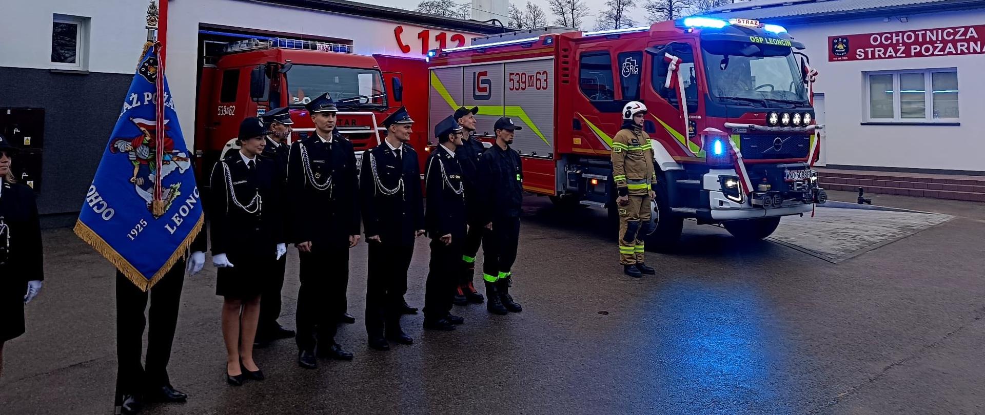 Strażacy OSP stoją na parkingu, jeden z nich po lewej stronie zdjęcia trzyma sztandar. . W tle dwa czerwone samochody strażackie oraz biały budynek z napisem „OCHOTNICZA STRAŻ POŻARNA”. 