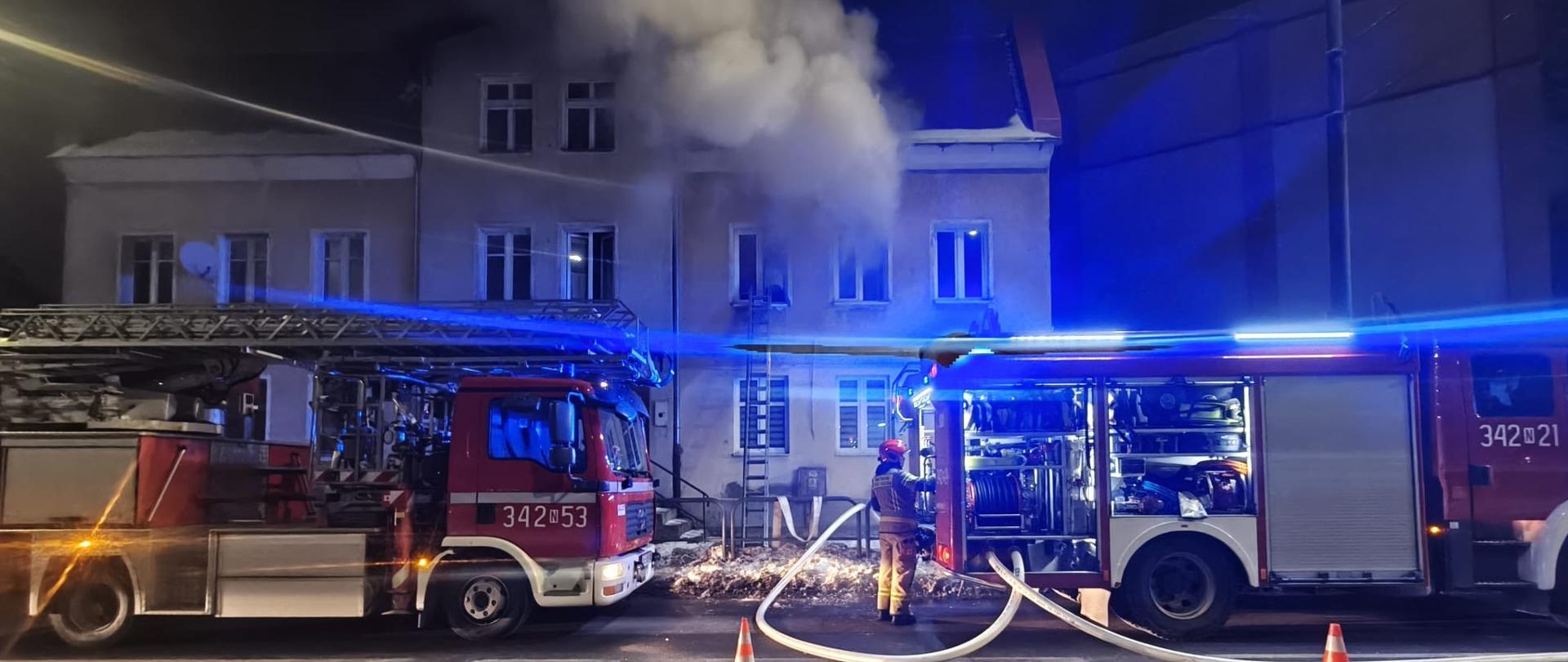 Strażacy pracują przy dwóch wozach pożarniczych stojących na ulicy, oświetlonych migającymi, niebieskimi światłami. Z jednego z mieszkań na wyższej kondygnacji wydobywa się gęsty dym, co wskazuje na pożar wewnątrz. Do budynku prowadzą rozwinięte węże, a teren zabezpieczono pachołkami drogowymi. Cała scena pokazuje intensywną, nocną interwencję służb ratowniczych.