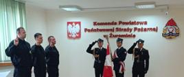 Na zdjęciu widzimy dalszy ciąg ceremonii ślubowania. Po lewej stronie znajdują się adepci, którzy mają podniesione dłonie w geście przysięgi. Na środku ściany widnieje napis "Komenda Powiatowa Państwowej Straży Pożarnej w Żurominie", co wskazuje na miejsce, w którym odbywa się ceremonia. Po obu stronach napisu widoczne są godła - polskie godło państwowe oraz godło straży pożarnej.