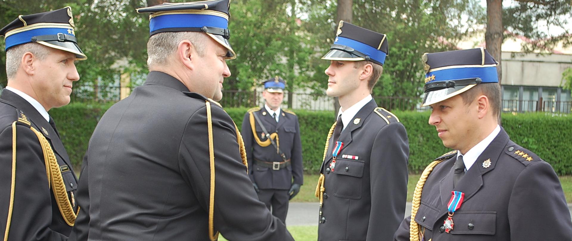 Zdjęcie przedstawia Zastępcę Małopolskiego Komendanta Wojewódzkiego bryg. Piotra Słowiaka podczas dekoracji odznaczeniami asp. Jakuba Pachoń, st. kpt Andrzeja Łotockiego. Z lewej strony asystujący Komendant Powiatowy. Na drugim planie stoi Dowódca uroczystości asp. sztab. Mariusz Masełko na tle zielonych krzewów.