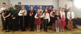 Na zdjęciu duża grupa ludzi. Są to uczniowie, którzy wystąpili podczas koncert klasy skrzypiec. Uczniowie trzymają skrzypce, jedna dziewczynka po prawej stronie trzyma gitarę. W tle za nimi niebieska tkanina, a na niej białe płatki śniegu. 