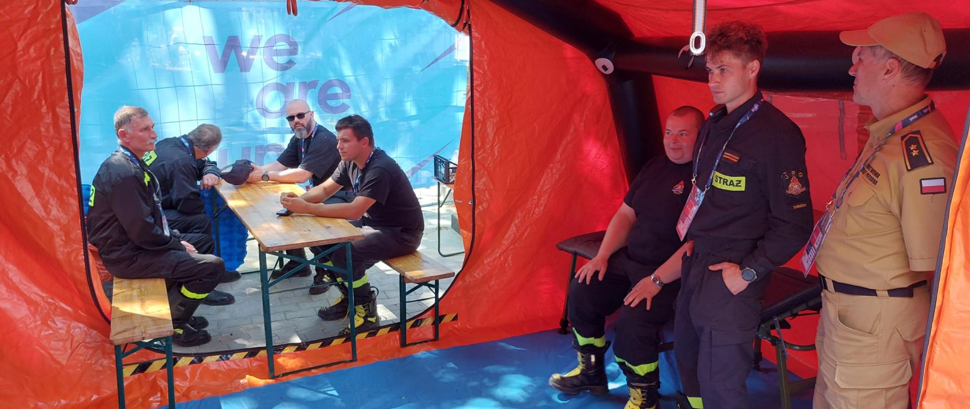 Zdjęcie przedstawia Sztab akcji zabezpieczenia III Igrzysk Europejskich. Strażacy znajdują się w pomarańczowym namiocie z niebieskim spodem.