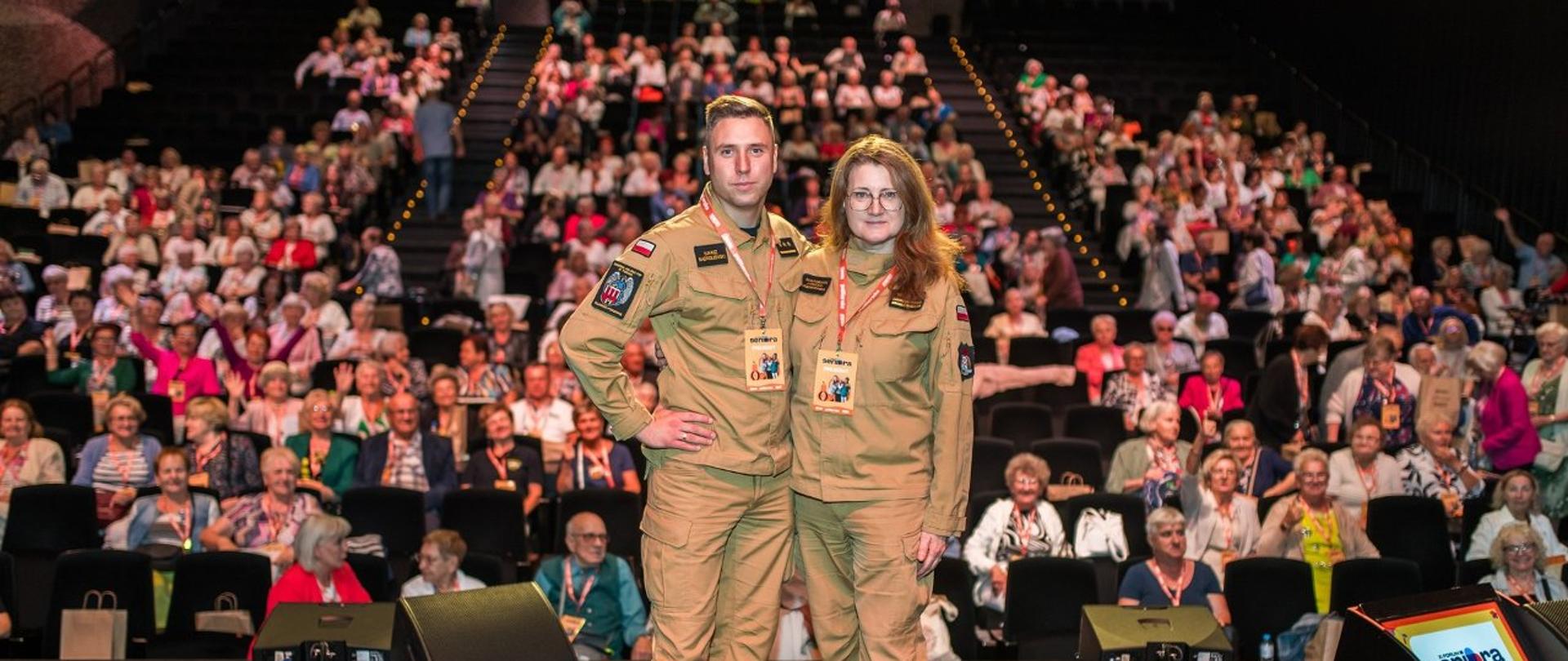 Dwoje strażaków w mundurach służbowych, stoją na scenie, za nimi tłum - kilkaset osób siedzących na widowni.