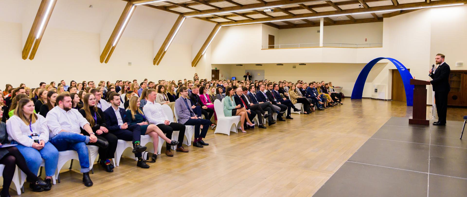 Przewodniczący Parlamentu Studentów RP mówi do mikrofonu, stojąc w wielkiej sali, przed nim ludzie siedzą na ustawionych w rzędy krzesłach.