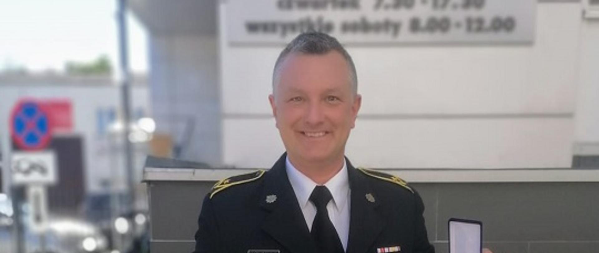 Zdjęcie przedstawia funkcjonariusza na tle budynku RCKiK w Bydgoszczy w umundurowaniu wyjściowym, trzymającego dyplom, legitymację oraz odznakę Honorowy Dawca Krwi - Zasłużony dla Zdrowia Narodu.