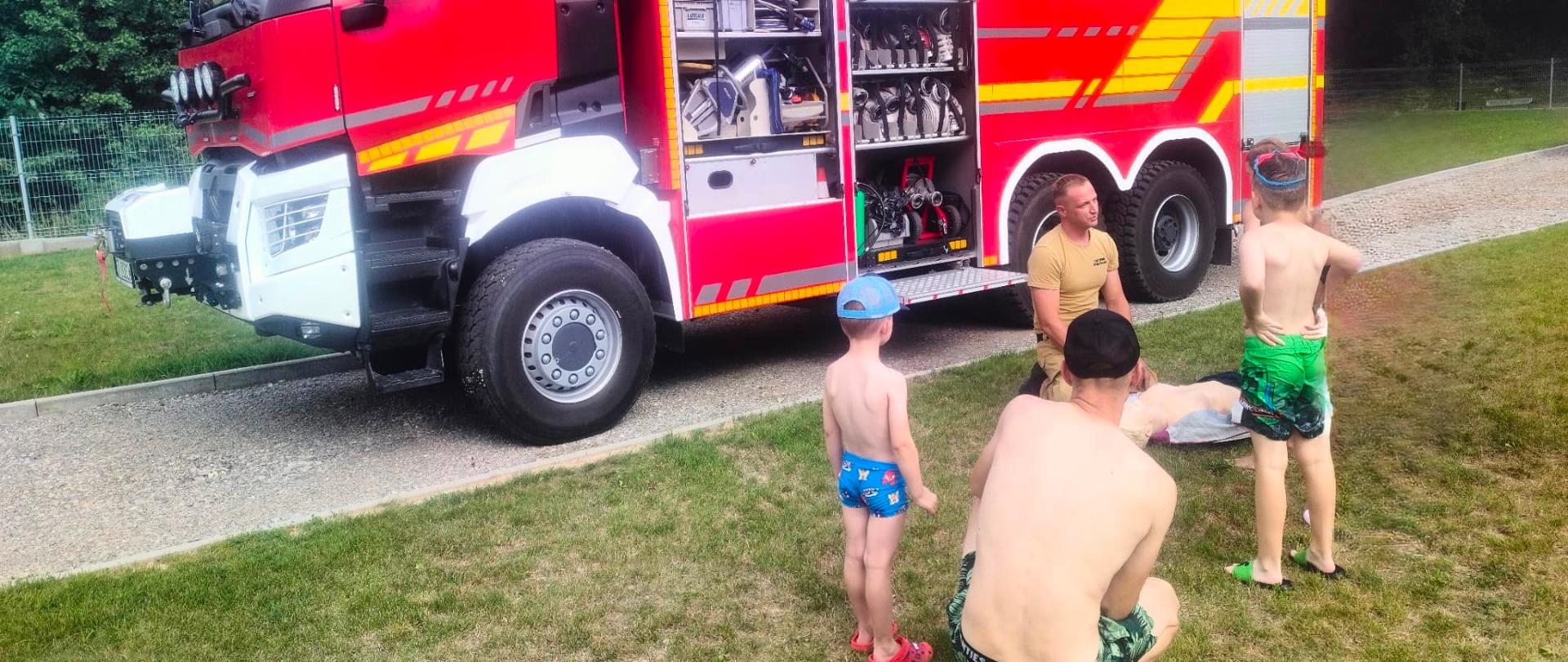 Strażak Państwowej Straży Pożarnej prowadzi pokaz pierwszej pomocy dla dzieci i dorosłych obok otwartego samochodu ratowniczo-gaśniczego. W tle widoczne są przedziały sprzętowe wozu strażackiego z wyposażeniem ratowniczym, a uczestnicy obserwują prezentację na trawniku.