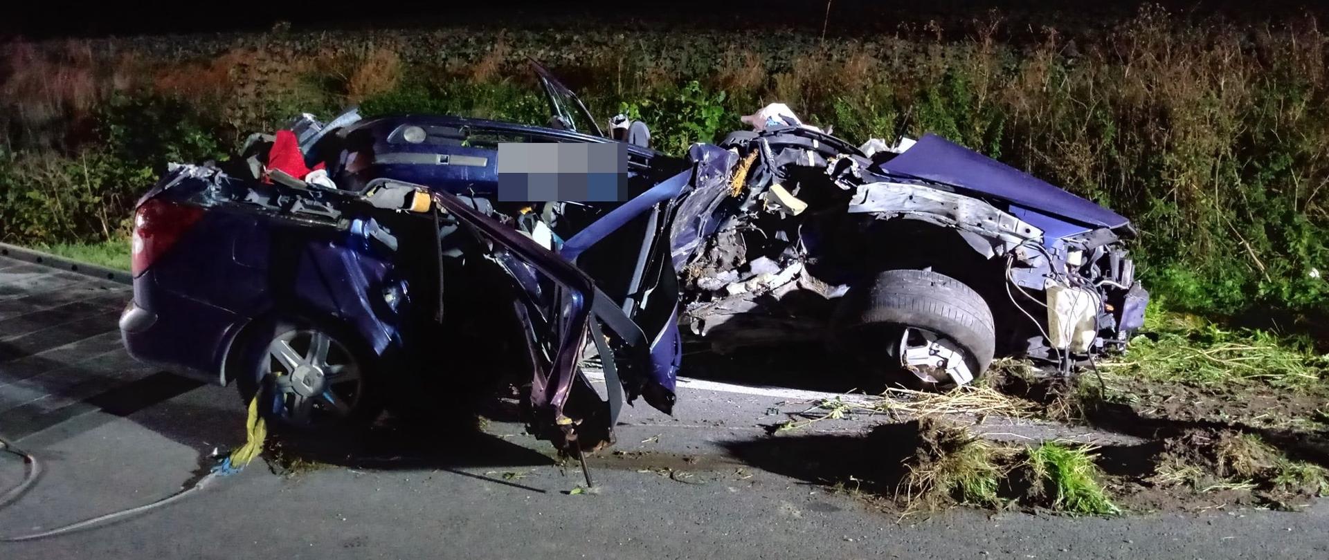 Częściowo na jezdni, częściowo na poboczu stoi mocno zniszczony i zniekształcony w wyniku uderzenia w drzewo wrak samochodu osobowego koloru niebieskiego. Pora nocna, za samochodem rozpościera się pole.