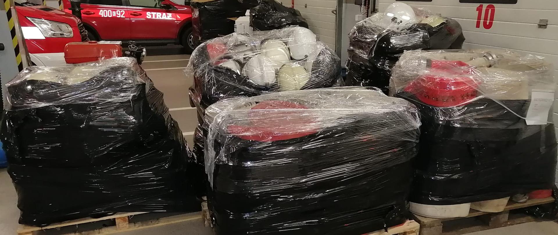 Zdjęcie w centralnej części ukazuje palety, na których znajduje się zabezpieczony folią sprzęt strażacki. Palety stoją na posadzce garażu. Na drugim planie widać samochód osobowy strażacki