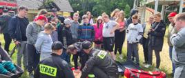 zdjęcie przedstawia grupę młodzieży i dzieci podczas pokazów strażackich z zakresu udzielania pierwszej pomocy. Strażacy w umundurowaniu koszarowym pokazują jak wykonywać resuscytację krążeniowo-oddechową. W trakcie wykonywania resuscytacji jedna z dziewczyn. Obok torba medyczna PSP R1. W oddali budynki i samochód strażacki.