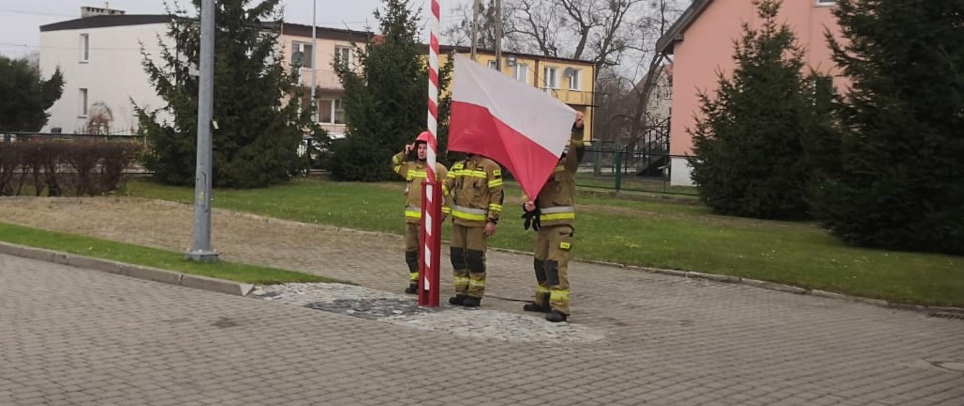 Strażacy stoją przy maszczie na placu i wciągają flage pańśtwową. Za nimi widać domy jednorodzinne.