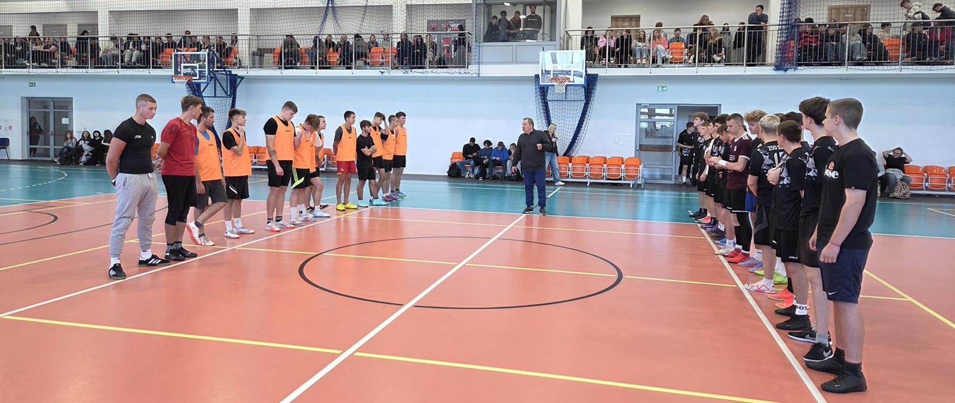 Dwie chłopięce drużyny stoją naprzeciwko siebie w hali sportowej, pomiędzy nimi stoi sędzia, w tle widać publiczność. 