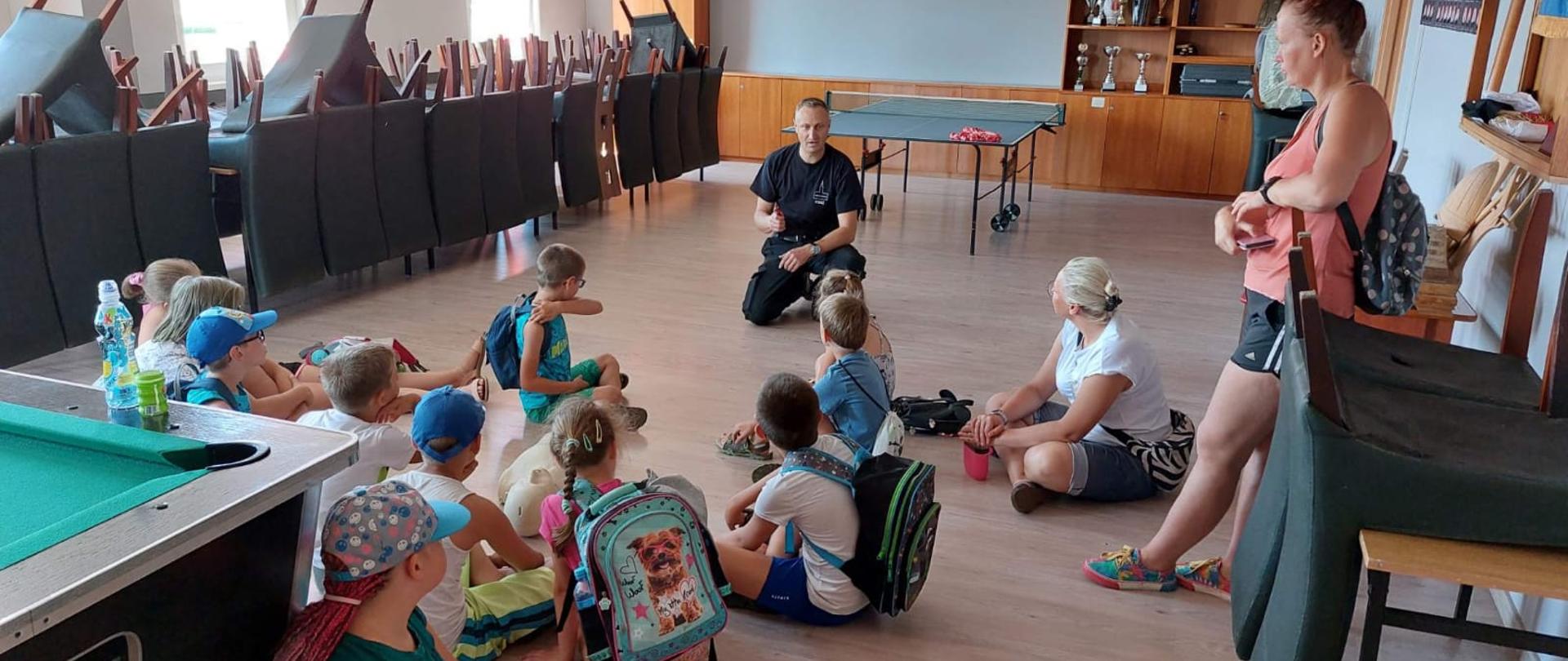 Na zdjęciu znajdują się dzieci, opiekunka i strażak. Dzieci siedzą na podłodze na świetlicy.