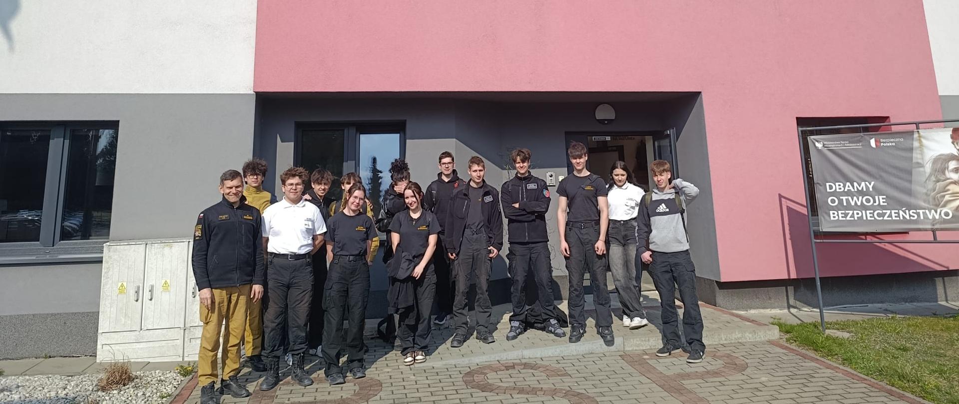 Uczniowie i strażak stoją na tle ściany budynku. Nad ich głowami napis KOMENDA MIEJSKA PAŃSTWOWEJ STRAŻY POŻARNEJ W ŻORACH. Po prawej stronie baner z napisem Dbamy o Twoje bezpieczeństwo. 
