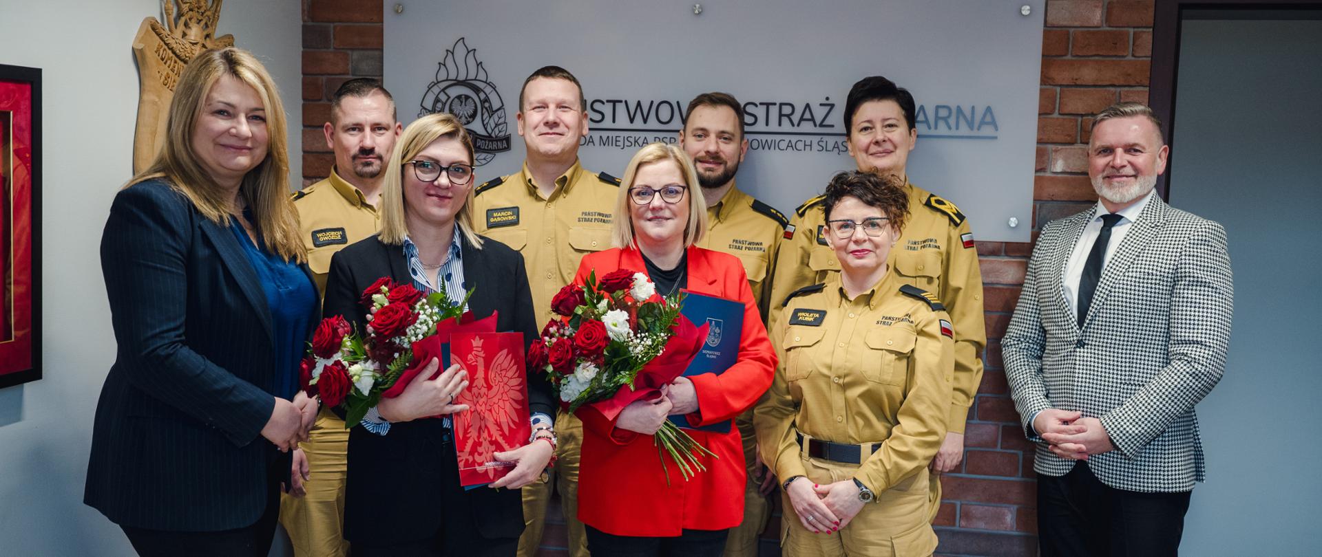 Grupowe zdjęcie w pomieszczeniu służbowym: na pierwszym planie stoi kilka osób, w środku dwie osoby trzymają bukiety czerwono-białych kwiatów i teczki. W tle na ceglanej ścianie widoczna jest tablica z napisem ‘Państwowa Straż Pożarna’.