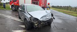 Samochód osobowy stoi na jezdni z uszkodzonym przodem. Z tyłu stoi samochód strażacki i stoją strażacy. 