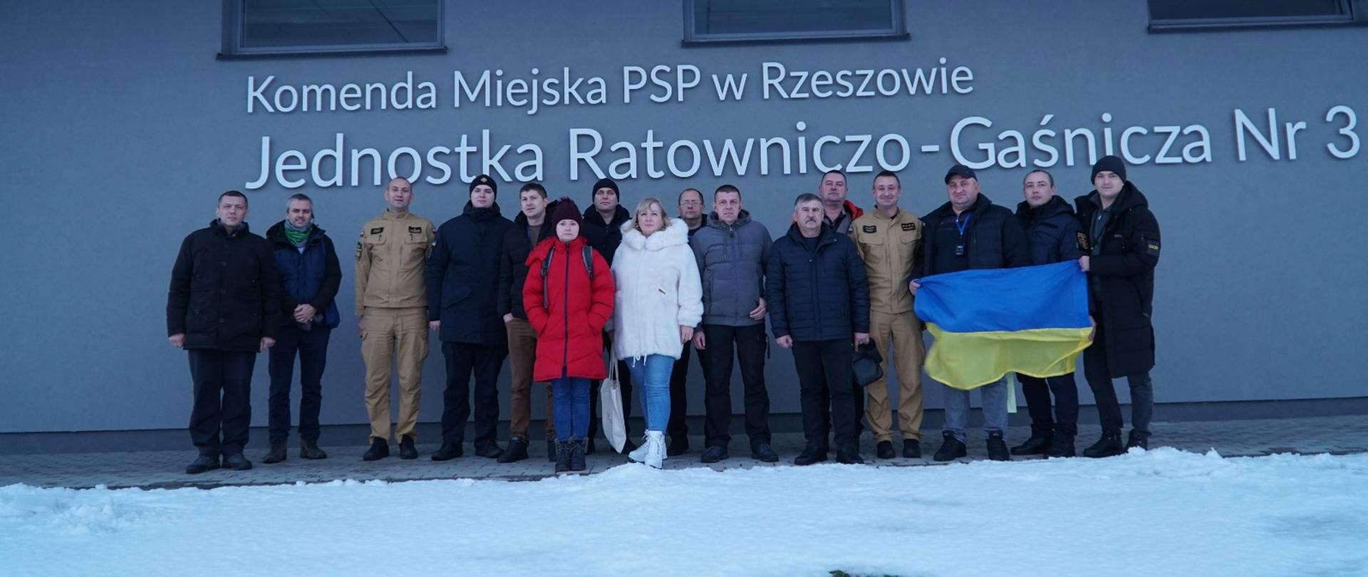 Zdjęcie przedstawia uczestników projektu „Pogromcy ognia. Wzmacnianie odporności ukraińskich hromad poprzez miejscowe ochotnicze organizacje ochrony ludności" wraz z dowództwem Jednostki Ratowniczo-Gaśniczej nr 3 w Rzeszowie. Zdjęcie zostało wykonane przed Jednostką Ratowniczo-Gaśniczą nr 3, jeden z uczestników trzyma w rękach flagę Ukrainy.