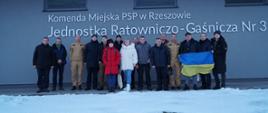 Zdjęcie przedstawia uczestników projektu „Pogromcy ognia. Wzmacnianie odporności ukraińskich hromad poprzez miejscowe ochotnicze organizacje ochrony ludności" wraz z dowództwem Jednostki Ratowniczo-Gaśniczej nr 3 w Rzeszowie. Zdjęcie zostało wykonane przed Jednostką Ratowniczo-Gaśniczą nr 3, jeden z uczestników trzyma w rękach flagę Ukrainy.