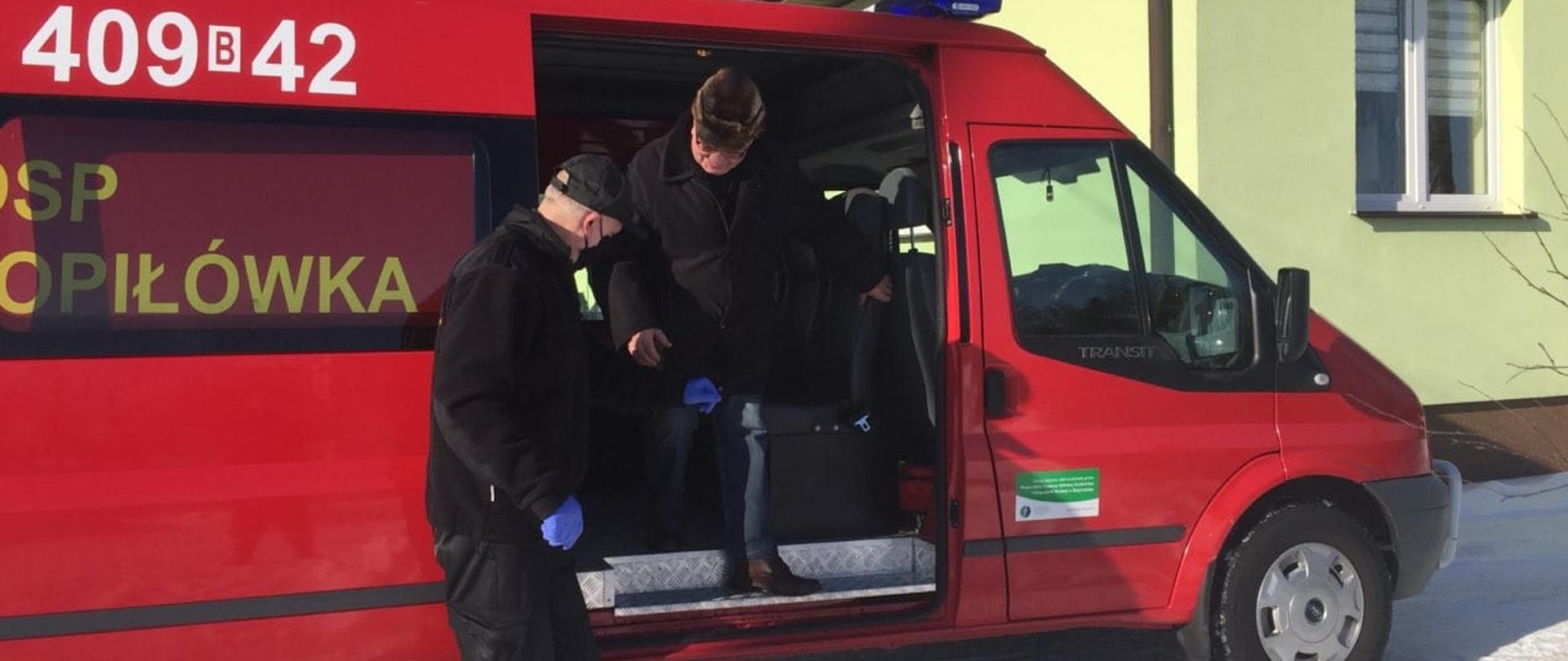 W tle widać samochód pożarniczy podczas dowozu osoby do punktu szczepień oraz strażaka pomagającemu starszej osobie w wyjściu z pojazdu. 