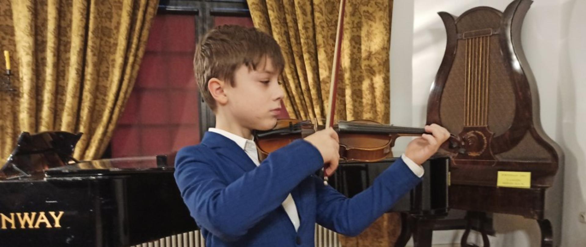 Zdjęcie kolorowe. Chłopiec w niebieskim garniturze gra na skrzypcach. Za nim stoi fortepian, z prawej strony stoi zabytkowy fortepian - lira.