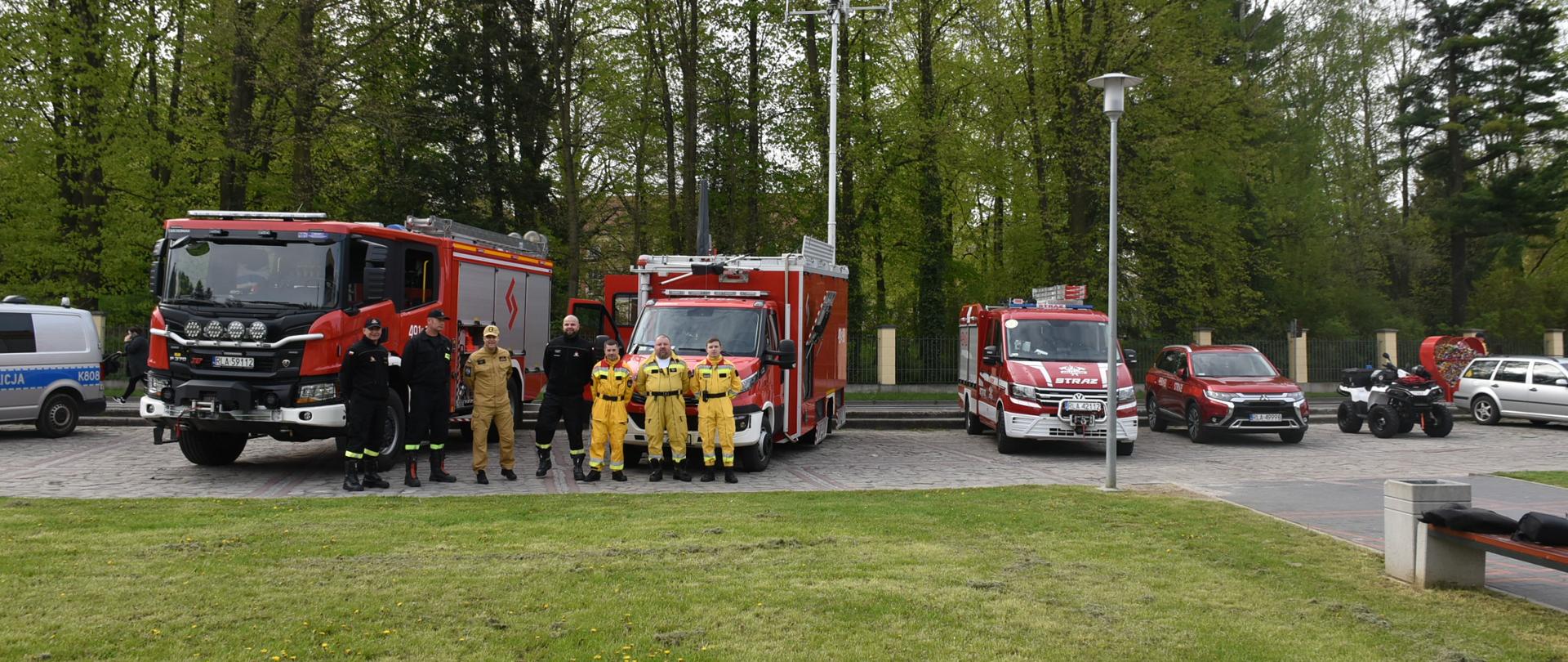 Zdjęcie przedstawia plac przed miejskim domem kultury na którym ustawione są pojazdy komendy powiatowej państwowej straży pożarnej w Łańcucie oraz ochotniczej straży pożarnej w Husowie przed którymi stoją strażacy tych jednostek. W tle park w Łańcucie.