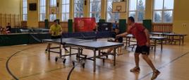 Dwóch mężczyzna w strojach sportowych gra w tenisa stołowego w sali gimnastycznej, w tle widoczne kolejne stoły do tenisa
