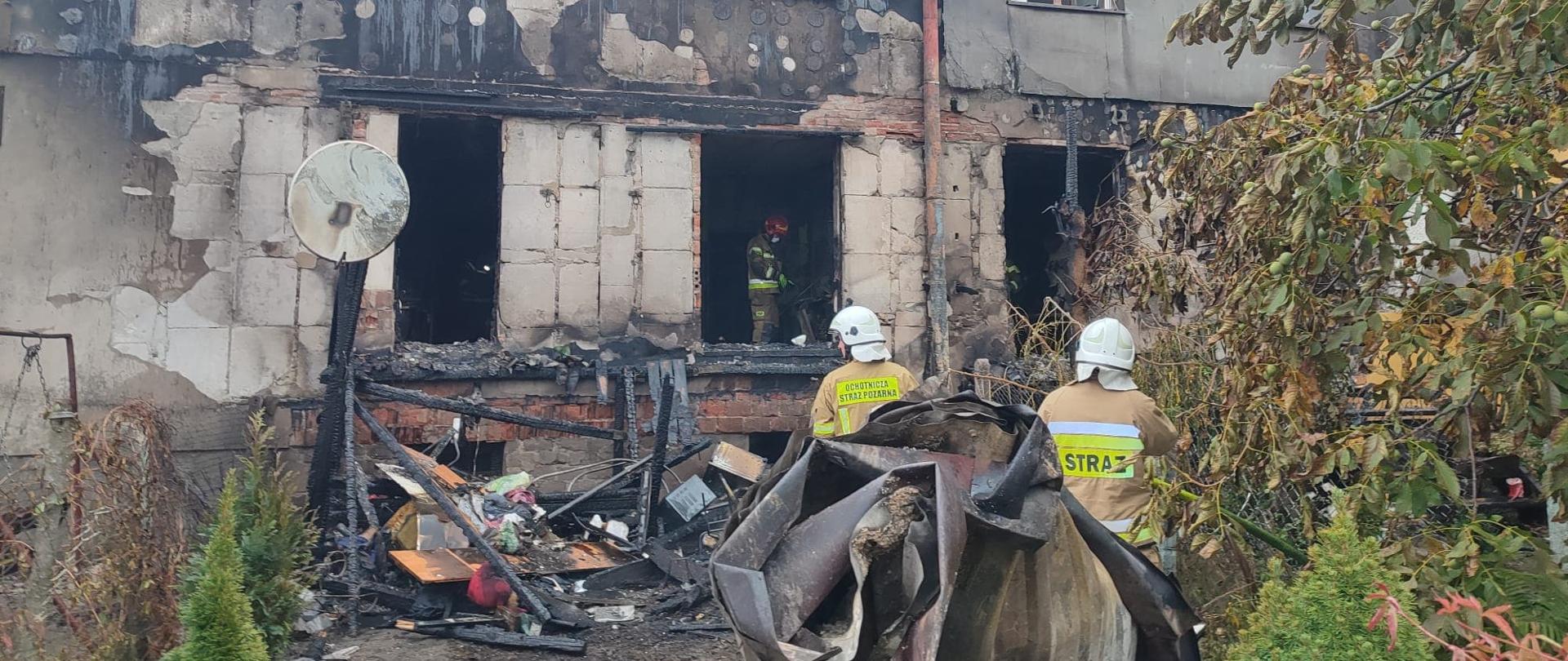Zniszczony przez pożar blok mieszkalny. Wśród pogorzeliska widać 3 strażaków w ubraniach specjalnych.
