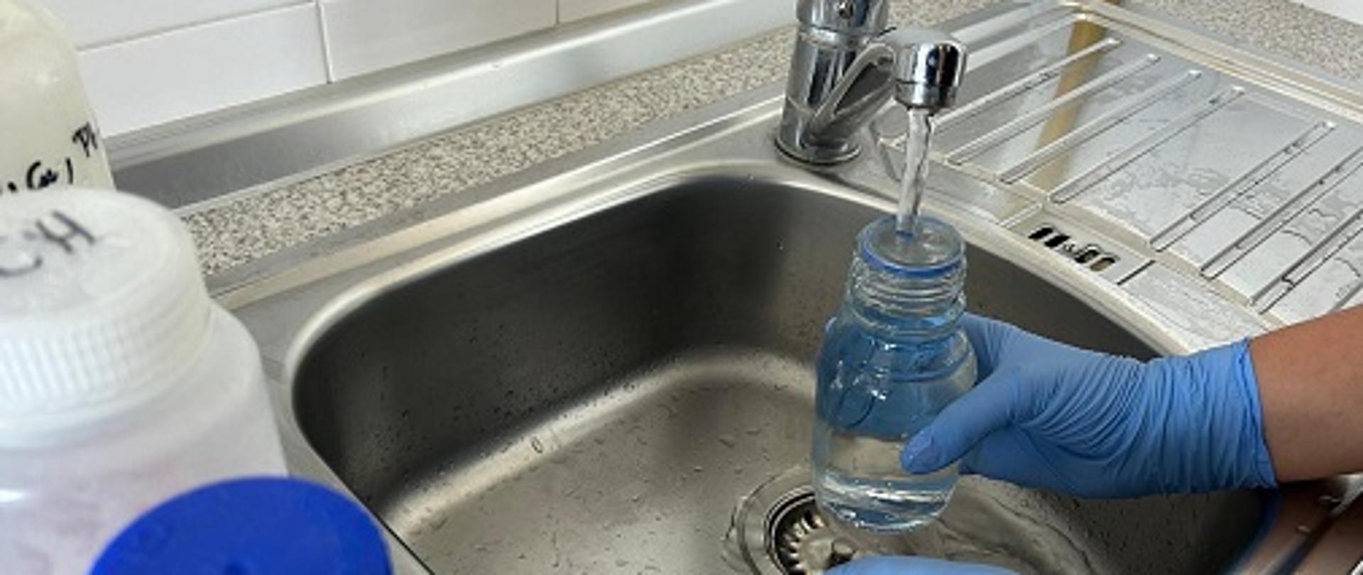 osoba w jednorazowych rękawicach ochronnych pobiera próbkę wody z kranu